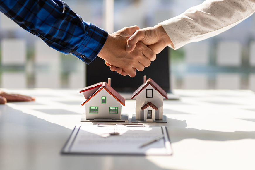 Two people shaking hands over some paperwork and two models of houses