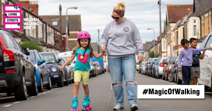 A woman walking down the middle of a street holding the hand of a young girl who is using roller skates. Logos for "Living Streets" and "National Walking Month". #MagicOfWalking